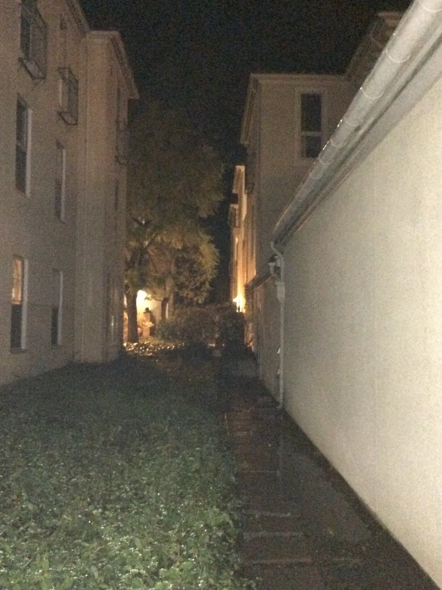 A narrow gap between two apartment blocks at night, one lit tree and a small figure at the far end.