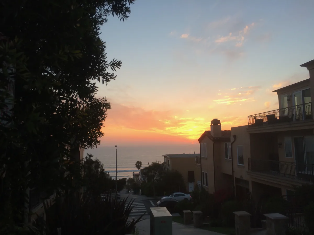 Sunset over the Pacific seen down a hillside street, the houses along it already in silhouette.