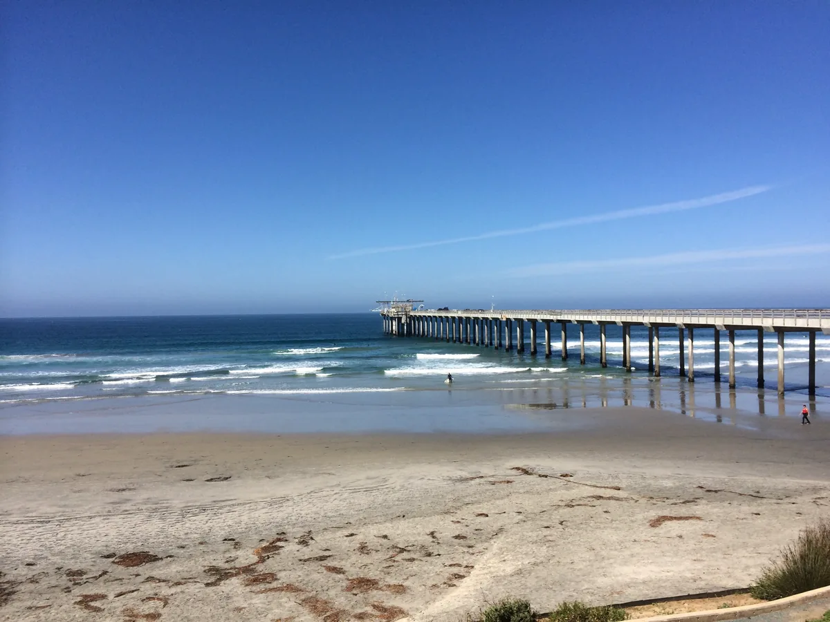 A long concrete research pier running out over the surf under a cloudless sky; one surfer, one walker.