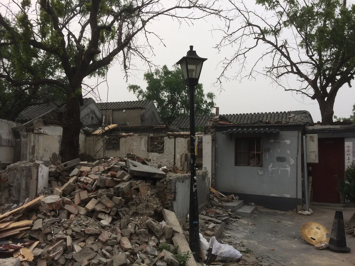 A hutong half demolished: brick rubble to the roofline, a lamp post still standing, one door still red.