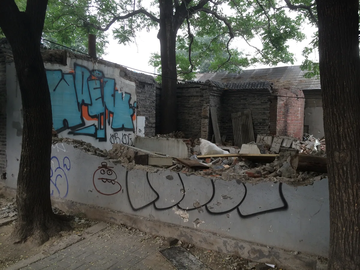 Graffiti on a low wall broken open, the courtyard behind it reduced to bricks and loose boards.