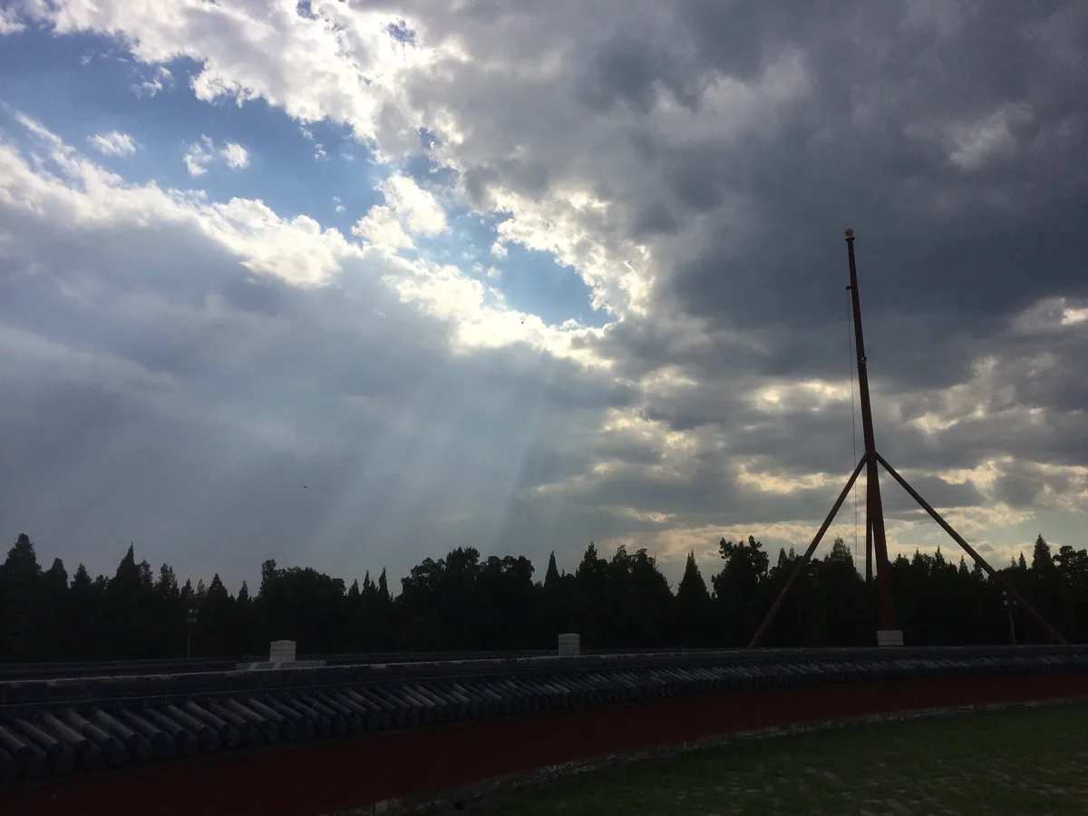 Sun breaking through heavy cloud over a tiled temple wall and cypresses, a tall mast at the right.