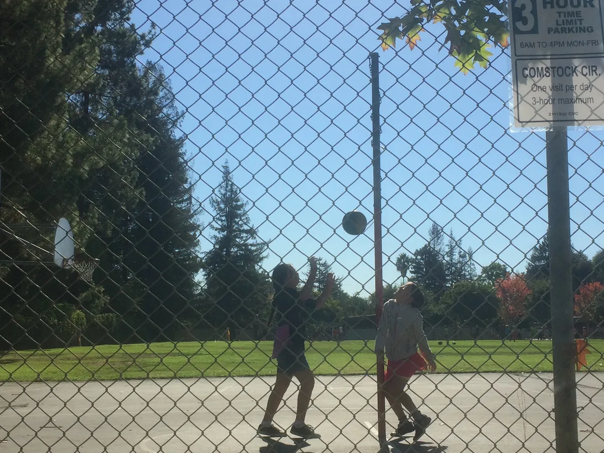Through a chain-link fence, two kids and a ball in mid-air on an otherwise empty court.