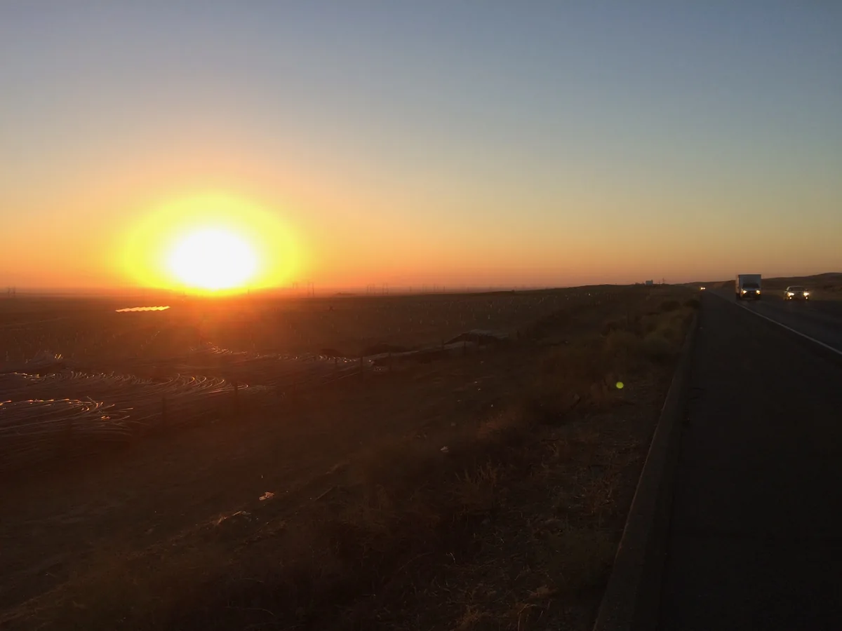 Sun on the horizon over flat farmland beside a freeway, coiled irrigation pipe stacked in the field.