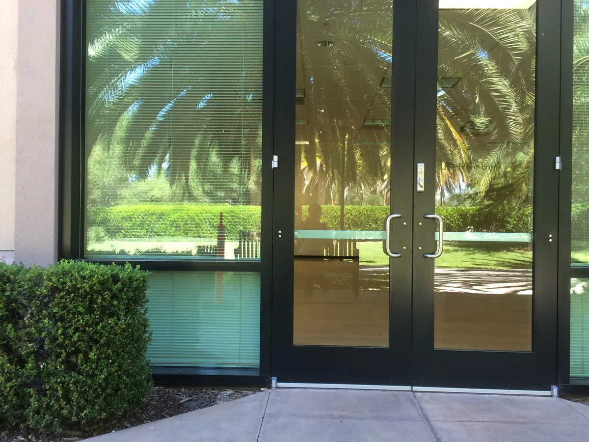 Glass office doors mirroring palms and lawn, the lit interior showing faintly through the reflection.