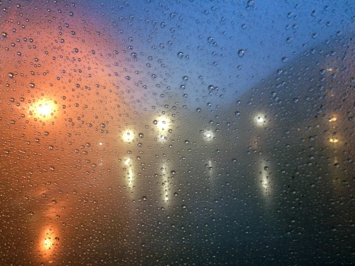 Rain beading on a window at dusk, the streetlights behind it dissolved into orange and blue smears.