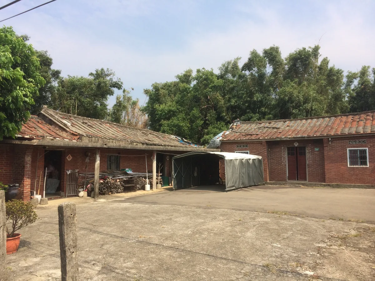 A red-brick courtyard farmhouse with a patched tile roof, firewood stacked and a tarp carport in the yard.
